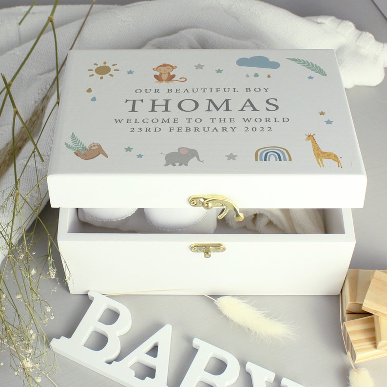 May include: A white wooden keepsake box with a gold clasp, open to reveal baby items. The lid is decorated with a sun, monkey, sloth, elephant, rainbow, giraffe, and text that reads "OUR BEAUTIFUL BOY THOMAS WELCOME TO THE WORLD 23RD FEBRUARY 2022."