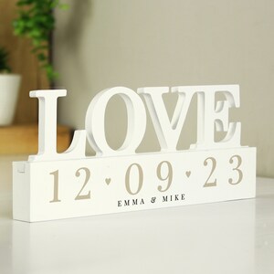 May include: White wooden sign with the word "LOVE" in large letters and the date "12.09.23" and the names "Emma & Mike" below.