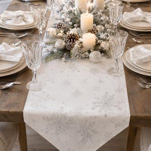May include: A white table runner featuring a silver snowflake pattern, displayed on a wooden dining table set with white plates, crystal glasses, silver cutlery, white napkins with silver rings, and a centrepiece of candles and ornaments.