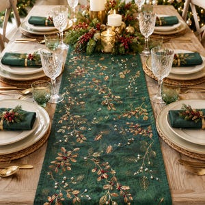 May include: A dark green table runner featuring an embroidered pattern of gold leaves, red berries, and small white beads. The runner is centred on a wooden dining table set with white plates, green napkins, gold cutlery, and crystal glassware.