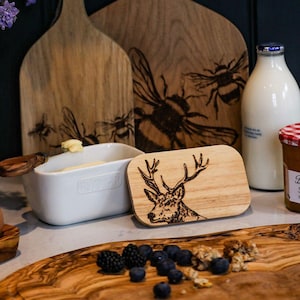 Op de afbeelding: Een witte keramische botervloot met een houten deksel met een lasergegraveerd ontwerp van een hert met geweien. Het deksel ligt op een houten snijplank met blauwe bessen en bramen.