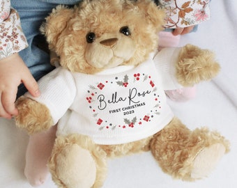May include: A tan plush teddy bear wearing a white knitted sweater. The sweater features a printed holiday wreath design with red berries, green holly leaves, and small gift boxes. The text reads Bella Rose FIRST CHRISTMAS 2023 in black ink.