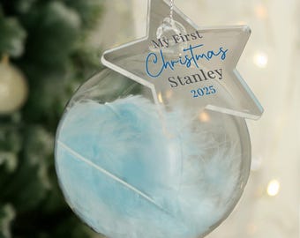 May include: Clear glass Christmas ornament with blue feathers inside. A clear star-shaped tag is attached, with the text "My First Christmas" and the name "Stanley" and the year "2025" in blue.