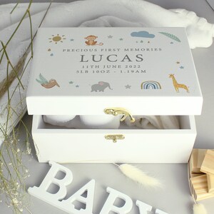 May include: White wooden keepsake box with a hinged lid, decorated with pastel illustrations of a sun, monkey, cloud, sloth, elephant, rainbow, and giraffe. The box is open, revealing a white blanket and baby shoes. The lid reads "PRECIOUS FIRST MEMORIES" and "LUCAS".
