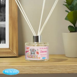 Op de afbeelding: Een heldere glazen diffuserfles met witte stokjes. De fles heeft een roze label met een cartoonbeer, bloemen en de tekst "To My Bestie Lauren Happy Birthday Love Amy x". De diffuser staat op een houten plank.