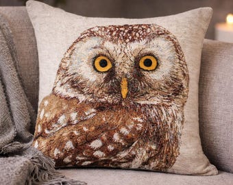 May include: A square cushion with an embroidered owl design. The owl has large yellow eyes, brown and white feathers, and a yellow beak. The cushion is set against a neutral backdrop, with a grey blanket and sofa visible.