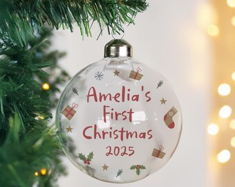 May include: A clear glass Christmas bauble with the text "Amelia's First Christmas 2025" in red. The bauble is decorated with small gift boxes, stars, snowflakes, and holly. It hangs from a silver metal cap on a Christmas tree.