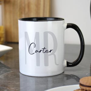 Pode incluir: Caneca de cerâmica branca com interior e alça pretos. A caneca apresenta o texto "MR Carter" em letras cinza e pretas. A caneca está sobre uma superfície cinza.