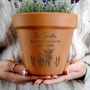Op de afbeelding: Een terracotta bloempot met de tekst "The Smiths, Rooted in Love, EST. 2026" en een illustratie van een Schotse hooglander. De pot is gevuld met lavendelplanten. De pot wordt door een persoon vastgehouden.