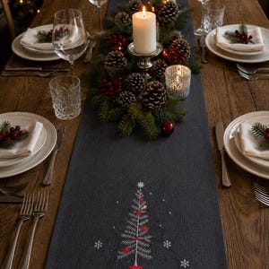 May include: A dark grey table runner featuring an embroidered white Christmas tree with red accents and small white snowflakes. The runner is placed on a wooden table set with white plates, clear glassware, and a centrepiece of pinecones and candles.