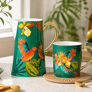 May include: A green ceramic jug and mug set with a parrot design. The jug has a gold handle and a white interior. The mug has a gold handle and sits on a woven coaster. The parrot design features orange, blue, and yellow colours.