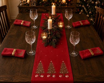 Puede incluir: Un camino de mesa rojo con árboles de Navidad marrones bordados y cajas de regalo blancas, sobre una mesa de madera con cuatro copas de vino de cristal, servilletas rojas con anillos dorados y un centro de velas, ramas y bayas.