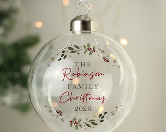 May include: Clear Christmas ornament with silver cap and white ribbon. The ornament has a festive design with the words "THE Robinson FAMILY Christmas 2023" in red script, surrounded by a wreath of green leaves, red berries, and gold snowflakes.