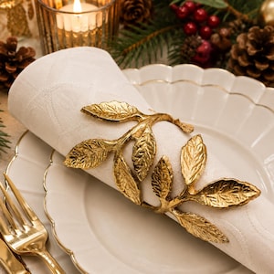 May include: A gold-toned metal napkin ring shaped like a leafy branch, featuring textured leaves and a stem, wrapped around a folded white fabric napkin with a subtle geometric pattern, resting on a white scalloped-edge ceramic plate.