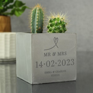 Puede incluir: Un macetero de hormigón gris con un diseño de corazón y el texto "MR & MRS 14.02.2023 EMMA & CHARLES FISHER" grabado en la parte delantera. Dos pequeñas plantas de cactus están creciendo en el macetero.