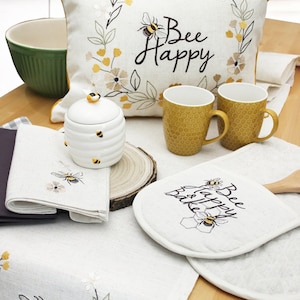 Puede incluir: Una mesa de cocina con un mantel de lino blanco, una almohada blanca con el texto "Bee Happy" bordado, dos tazas amarillas, un tarro de miel de cerámica blanca con abejas y dos agarraderas blancas con el texto "Bee Happy & Bake" bordado.