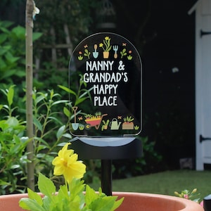 Può includere: Un paletto da giardino in metallo nero con un cartello in acrilico trasparente che recita "Nanny & Grandad's Happy Place". Il cartello è decorato con attrezzi da giardinaggio e piante colorate.
