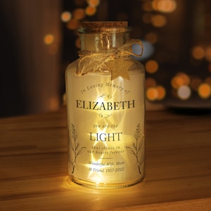May include: A clear glass jar with a cork lid and a white label. The label has a floral design and the text "In Loving Memory of ELIZABETH you are the LIGHT that shines in our hearts forever Wonderful Wife, Mum & Friend. 1957-2022".