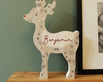 May include: A light-colored wooden reindeer figurine featuring a red nose and decorative patterns of stars, snowflakes, and pine needles. The name Benjamin is printed in red script across the side, with the year 2023 printed in black below it.
