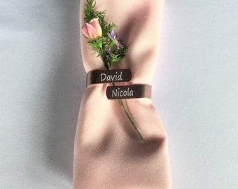 May include: A pink napkin with a flower and two copper napkin rings engraved with the names "David" and "Nicola".