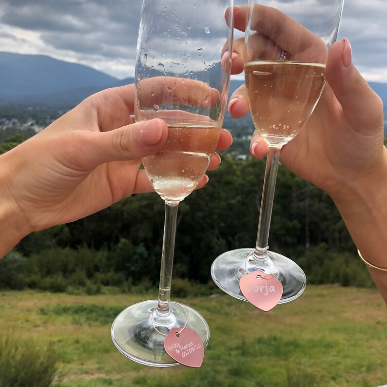 May include: Two champagne flutes with heart-shaped charms attached to the stems. The charms are engraved with the names "Krista & Marcus" and "Jorja".