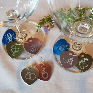 May include: Set of 10 heart-shaped wine glass charms with various designs, including a cake, balloons, a baby rattle, a bride and groom, and footprints. The charms are silver, gold, and copper.