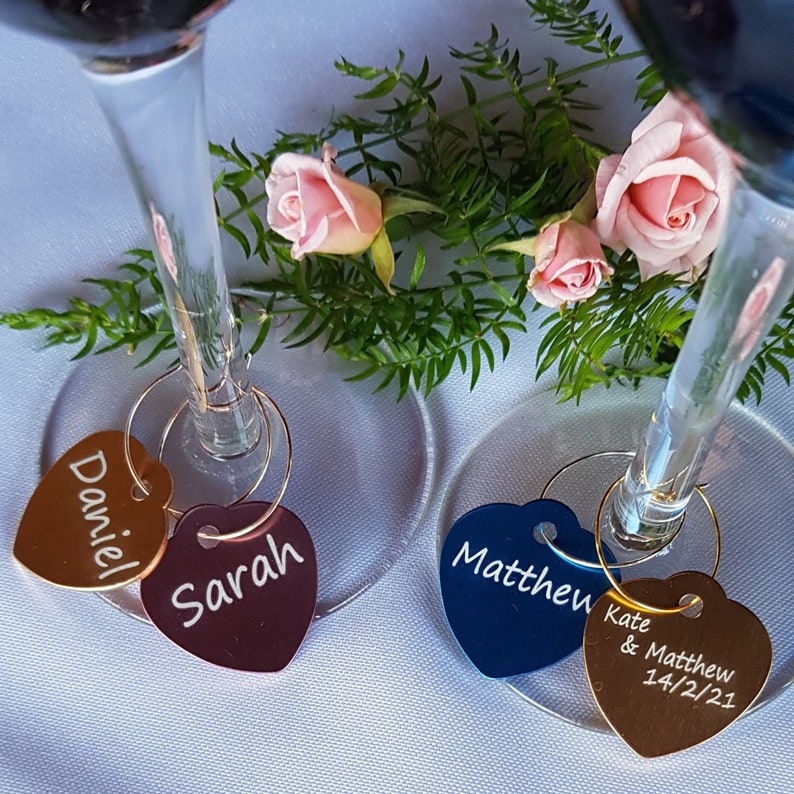 May include: Two wine glass charms with heart-shaped tags. One charm is gold with the name "Daniel" on it. The other charm is burgundy with the name "Sarah" on it. A third charm is blue with the name "Matthew" on it. The fourth charm is gold with the text "Kate & Matthew 14/2/21" on it.