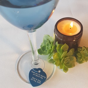 May include: A blue glass wine glass with a heart-shaped tag that says "Thank you for sharing our special day Steve & Amy 22 May 2021". The glass is on a white tablecloth with a small candle and succulents.