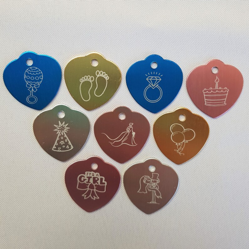 May include: Ten heart-shaped metal tags with various engraved designs, including a baby rattle, footprints, a wedding ring, a birthday cake, a party hat, a bride and groom, balloons, a bow with the text "It's a Girl", and a bride and groom holding hands.