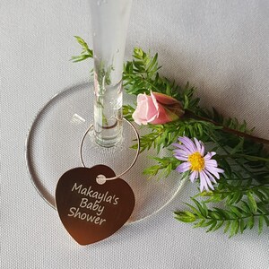 May include: A heart-shaped metal wine glass charm with the text "Makayla's Baby Shower" engraved on it. The charm is attached to a glass of water with a silver ring.