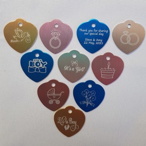 May include: Assortment of ten heart-shaped metal tags with various engraved designs. The tags are in shades of gold, silver, blue, pink, and green. The designs include a stork carrying a baby, a diamond ring, two wedding rings, a baby carriage, a cake, balloons, and the words "It's a Boy" and "It's a Girl!"