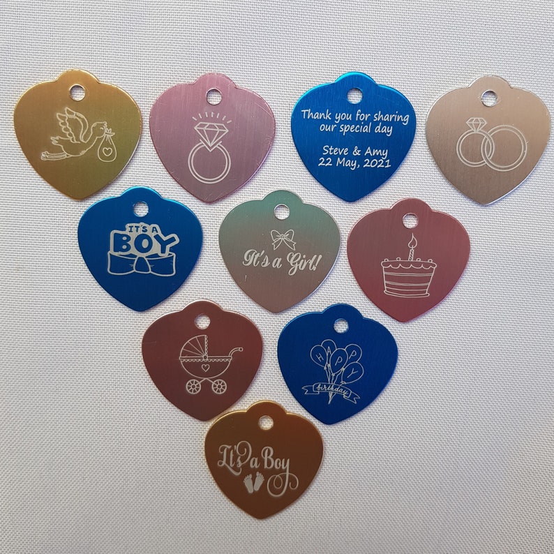 May include: Assortment of ten heart-shaped metal tags with various engraved designs. The tags are in shades of gold, silver, blue, pink, and green. The designs include a stork carrying a baby, a diamond ring, two wedding rings, a baby carriage, a cake, balloons, and the words "It's a Boy" and "It's a Girl!"
