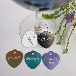 May include: Five heart-shaped metal wine glass charms with different names engraved on them. The charms are in various colors, including white, black, gold, teal, and purple.