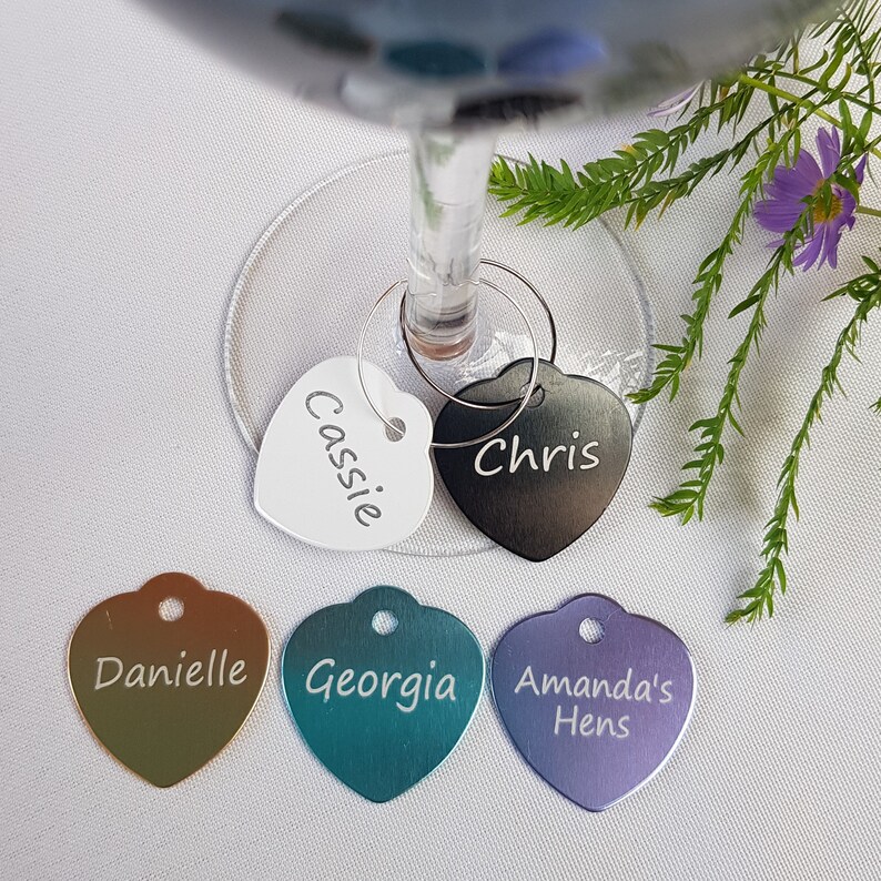 May include: Five heart-shaped metal wine glass charms with different names engraved on them. The charms are in various colors, including white, black, gold, teal, and purple.