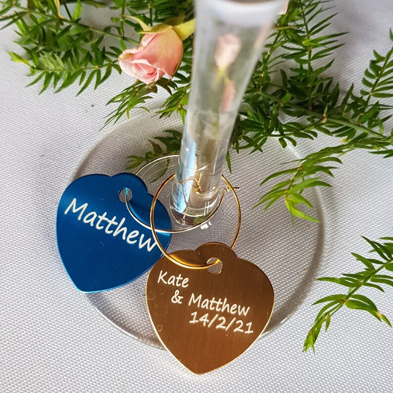 May include: Two heart-shaped wine glass charms, one blue and one gold. The blue charm says "Matthew" and the gold charm says "Kate & Matthew 14/2/21".