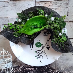 Pode incluir: Um prato de cer&acirc;mica branco com um chap&eacute;u preto e flores verdes no topo. Um rosto de mulher &eacute; pintado no prato com l&aacute;bios verdes e uma flor verde atr&aacute;s da cabe&ccedil;a.