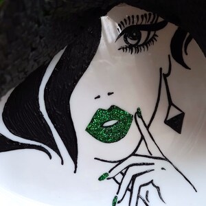 Pode incluir: Uma ilustra&ccedil;&atilde;o em preto e branco de um rosto de mulher com l&aacute;bios e esmalte de unha de glitter verde. A mulher est&aacute; usando um chap&eacute;u e tem a m&atilde;o no queixo.