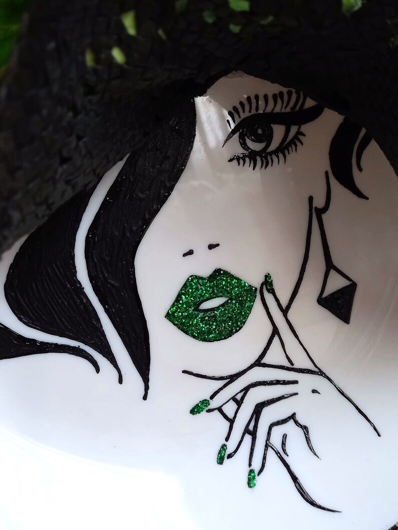 Pode incluir: Uma ilustra&ccedil;&atilde;o em preto e branco de um rosto de mulher com l&aacute;bios e esmalte de unha de glitter verde. A mulher est&aacute; usando um chap&eacute;u e tem a m&atilde;o no queixo.