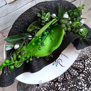 Pode incluir: Um chap&eacute;u preto tecido com enfeites florais verdes e brancos. O chap&eacute;u est&aacute; sentado em um prato branco com um design preto e branco.