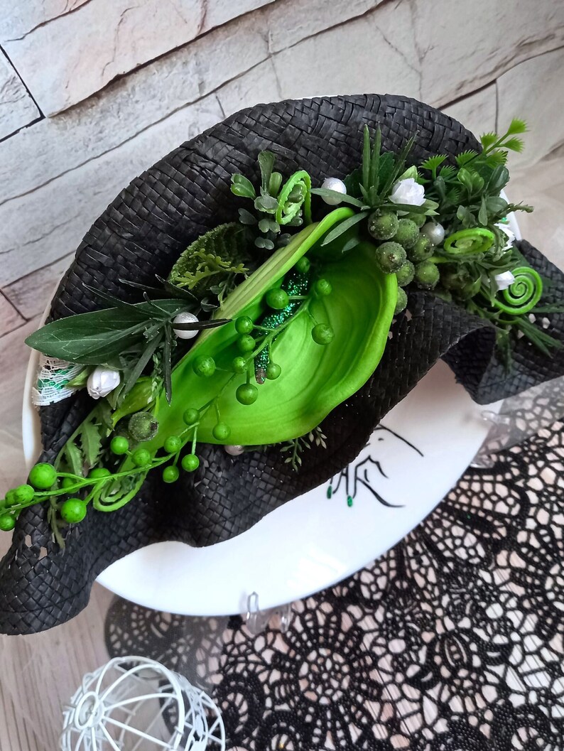 Pode incluir: Um chap&eacute;u preto tecido com enfeites florais verdes e brancos. O chap&eacute;u est&aacute; sentado em um prato branco com um design preto e branco.