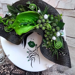 Pode incluir: Um prato de cer&acirc;mica branco com uma ilustra&ccedil;&atilde;o em preto e branco de um rosto de mulher com l&aacute;bios verdes. O prato &eacute; decorado com um chap&eacute;u tecido preto adornado com folhas e flores verdes.