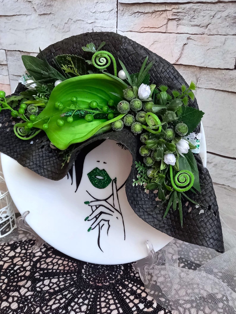 Pode incluir: Um prato de cer&acirc;mica branco com uma ilustra&ccedil;&atilde;o em preto e branco de um rosto de mulher com l&aacute;bios verdes. O prato &eacute; decorado com um chap&eacute;u tecido preto adornado com folhas e flores verdes.