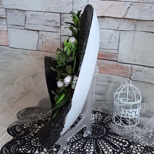 Pode incluir: Um chap&eacute;u tecido preto e branco com uma base branca e um arranjo floral verde e branco. O chap&eacute;u est&aacute; em um suporte de exibi&ccedil;&atilde;o branco.
