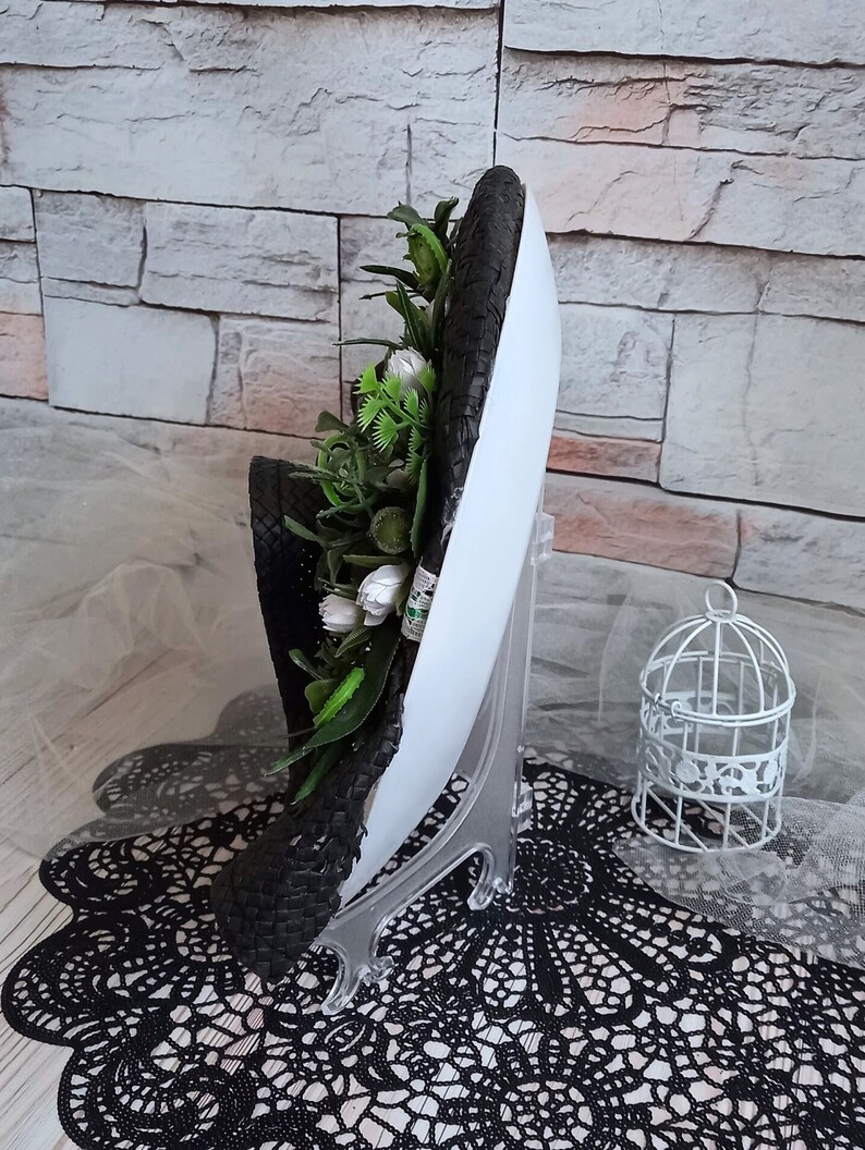 Pode incluir: Um chap&eacute;u tecido preto e branco com uma base branca e um arranjo floral verde e branco. O chap&eacute;u est&aacute; em um suporte de exibi&ccedil;&atilde;o branco.