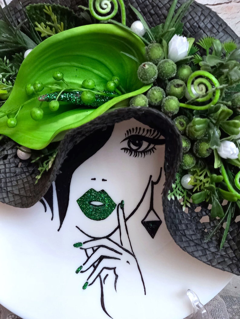 Pode incluir: Um chap&eacute;u preto e verde com uma flor verde e um rosto de mulher com l&aacute;bios verdes e esmalte de unha verde. O rosto da mulher &eacute; desenhado em preto e branco.