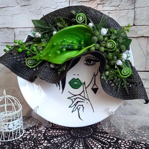 Pode incluir: Um prato de cer&acirc;mica branco com um design de chap&eacute;u preto e verde. O chap&eacute;u &eacute; decorado com folhas e flores verdes. &Eacute; poss&iacute;vel ver o rosto de uma mulher no prato, com l&aacute;bios verdes e esmalte de unhas verde.