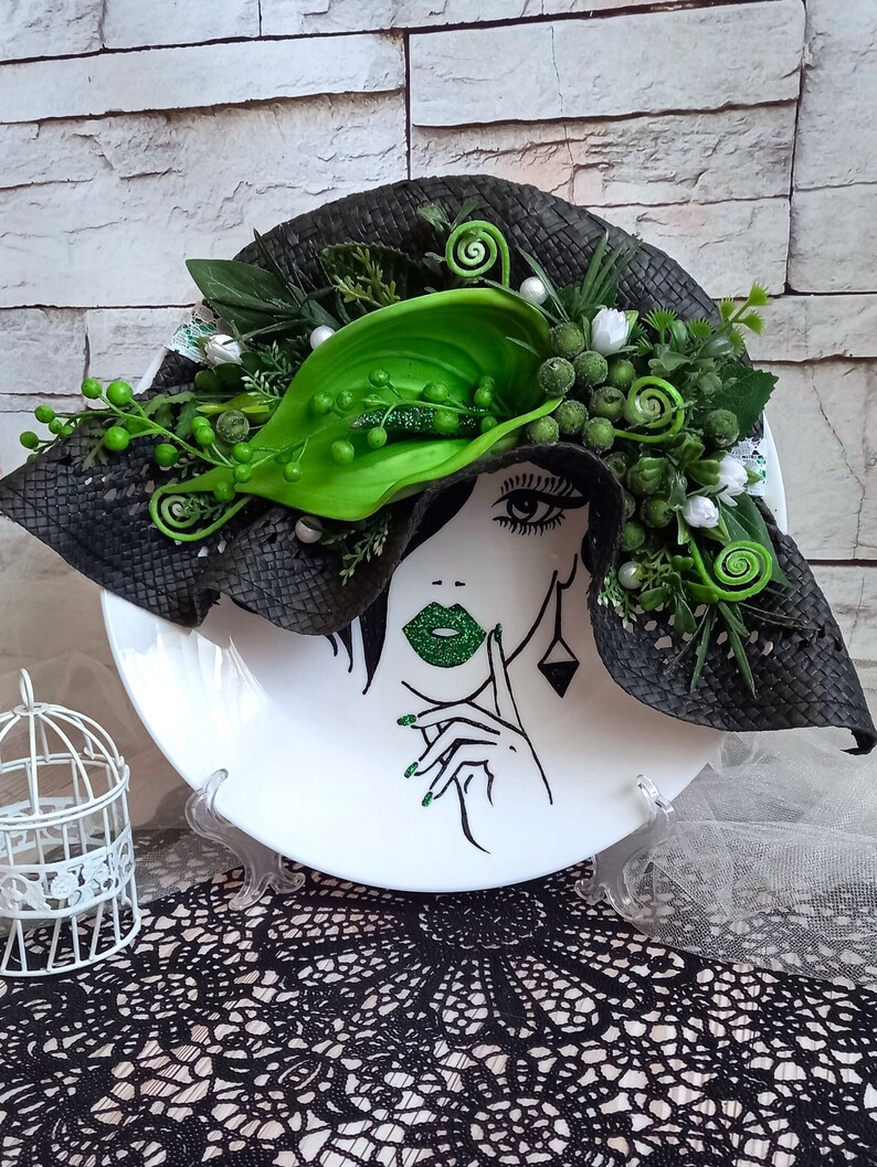 Pode incluir: Um prato de cer&acirc;mica branco com um design de chap&eacute;u preto e verde. O chap&eacute;u &eacute; decorado com folhas e flores verdes. &Eacute; poss&iacute;vel ver o rosto de uma mulher no prato, com l&aacute;bios verdes e esmalte de unhas verde.
