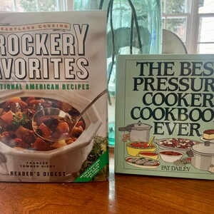 May include: Two cookbooks on a wooden table. The cookbook on the left is titled "Crocker Favorites" with the subtitle "Traditional American Recipes" and features a photo of a crock pot with a ladle. The cookbook on the right is titled "The Best Pressure Cooker Cookbook Ever" and features a photo of a pressure cooker and other cooking items.