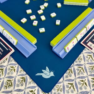 May include: A blue Mahjong game set with green and blue accents. The set includes a blue playing surface with a bird logo, tiles, and racks. The game is set up on a floral patterned tablecloth.