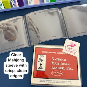 May include: A clear plastic protective sleeve for a 2024 National Mah Jongg League official hands and rules card, featuring a red cover with white text, alongside a light purple tile rack holding various colorful mahjong tiles with symbols.